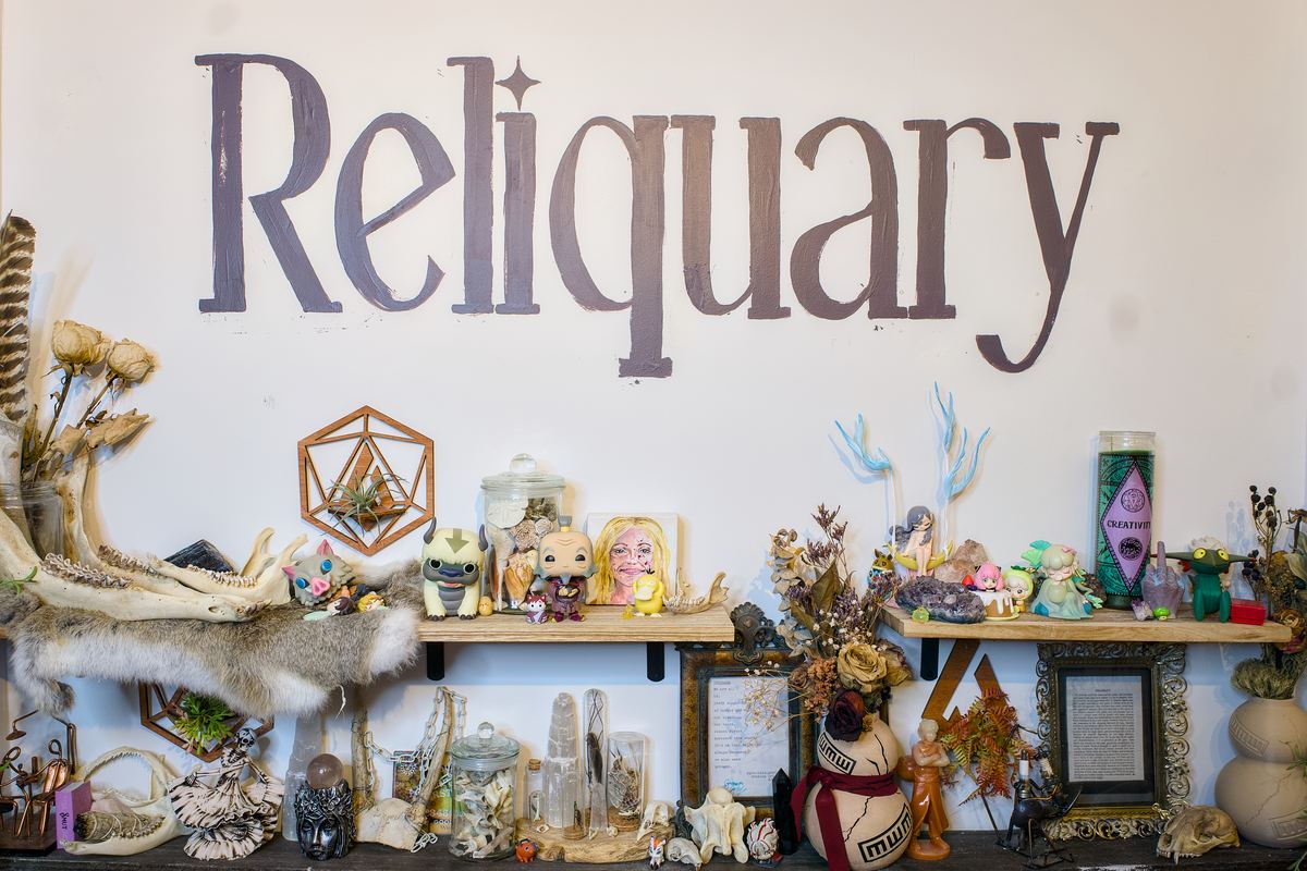 The Reliquary altar wall — figurines, dried flowers, crystals, skulls, and small relics arranged on shelves beneath the hand-painted wordmark.