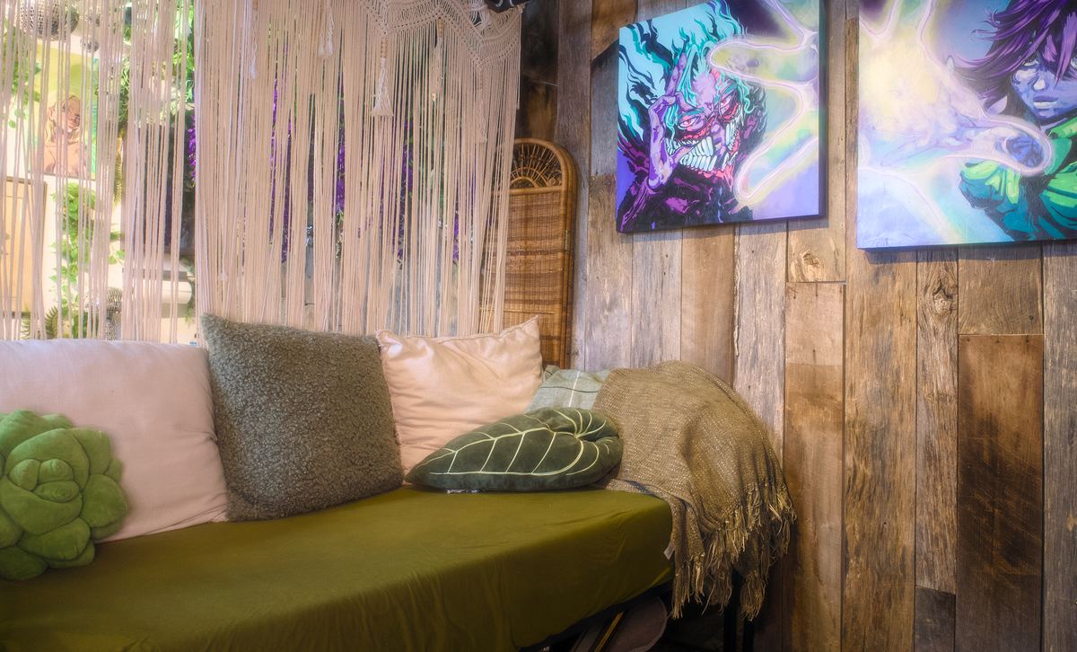 The studio's lounge corner — a green velvet day bed with cushions, a macrame curtain, and two demon-faced anime paintings on a reclaimed-wood wall.