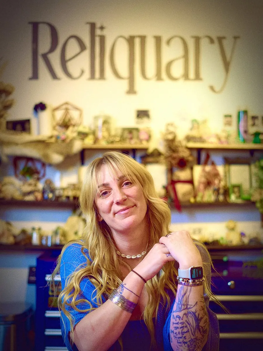 Sadie smiling, chin on her tattooed hands, under the hand-painted Reliquary wordmark and shelves of relics