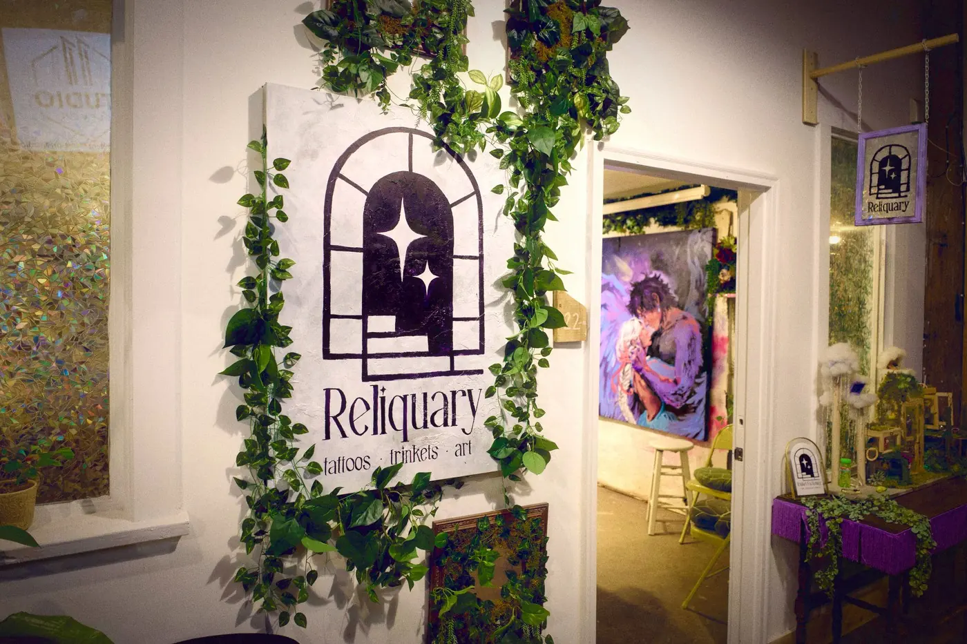 The Reliquary sign beside the studio door: the arched-window mark and "tattoos · trinkets · art", framed in trailing pothos, a painting glimpsed through the open door.