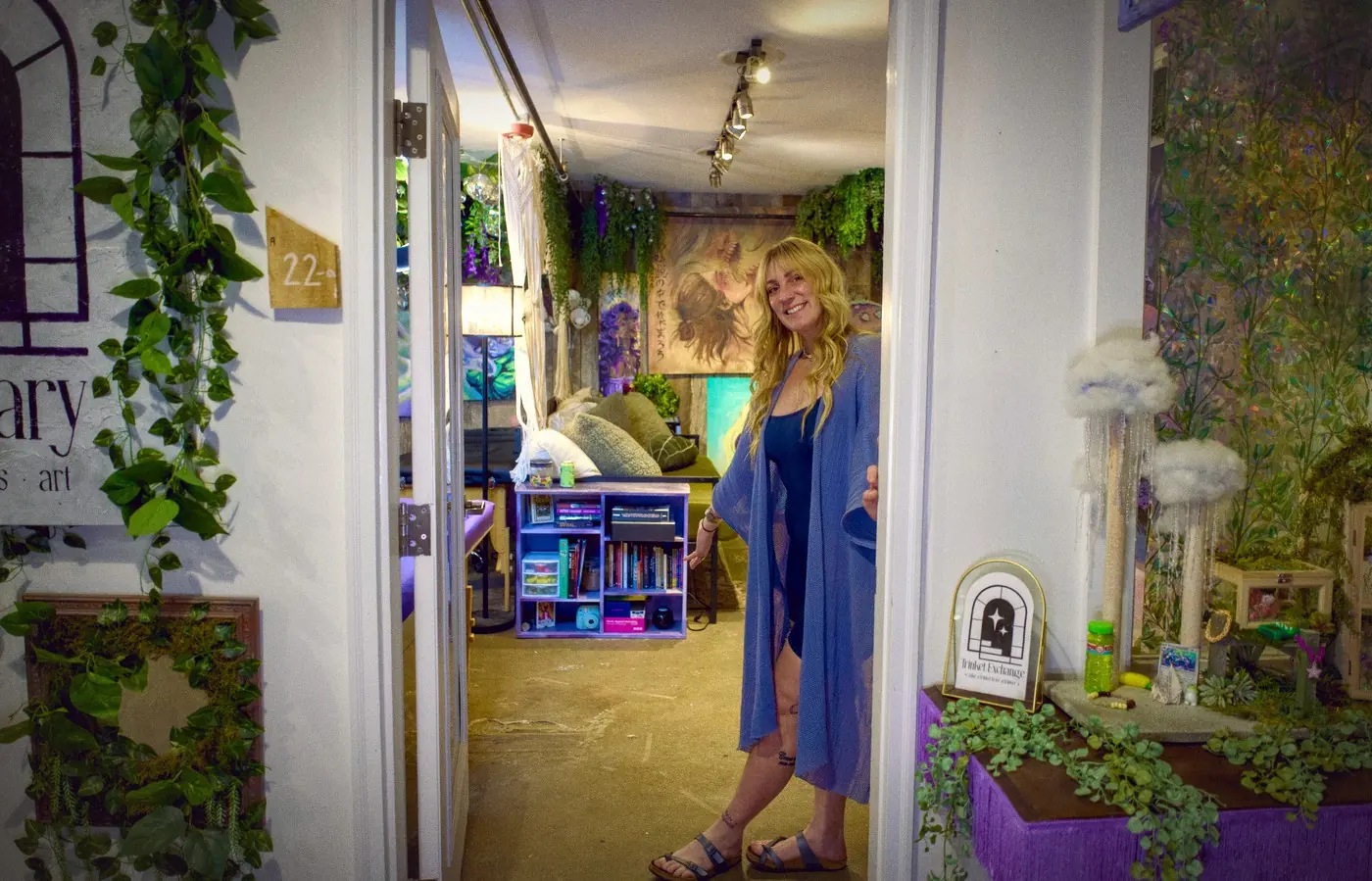 Sadie in the open door of Suite 22A, the studio behind her: plants, paintings and a lounge, the Trinket Exchange on a purple stand beside the door.