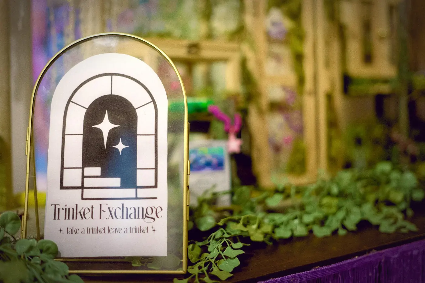 The Trinket Exchange sign in a brass arch, "take a trinket, leave a trinket", in front of the moss-lined cabinets.
