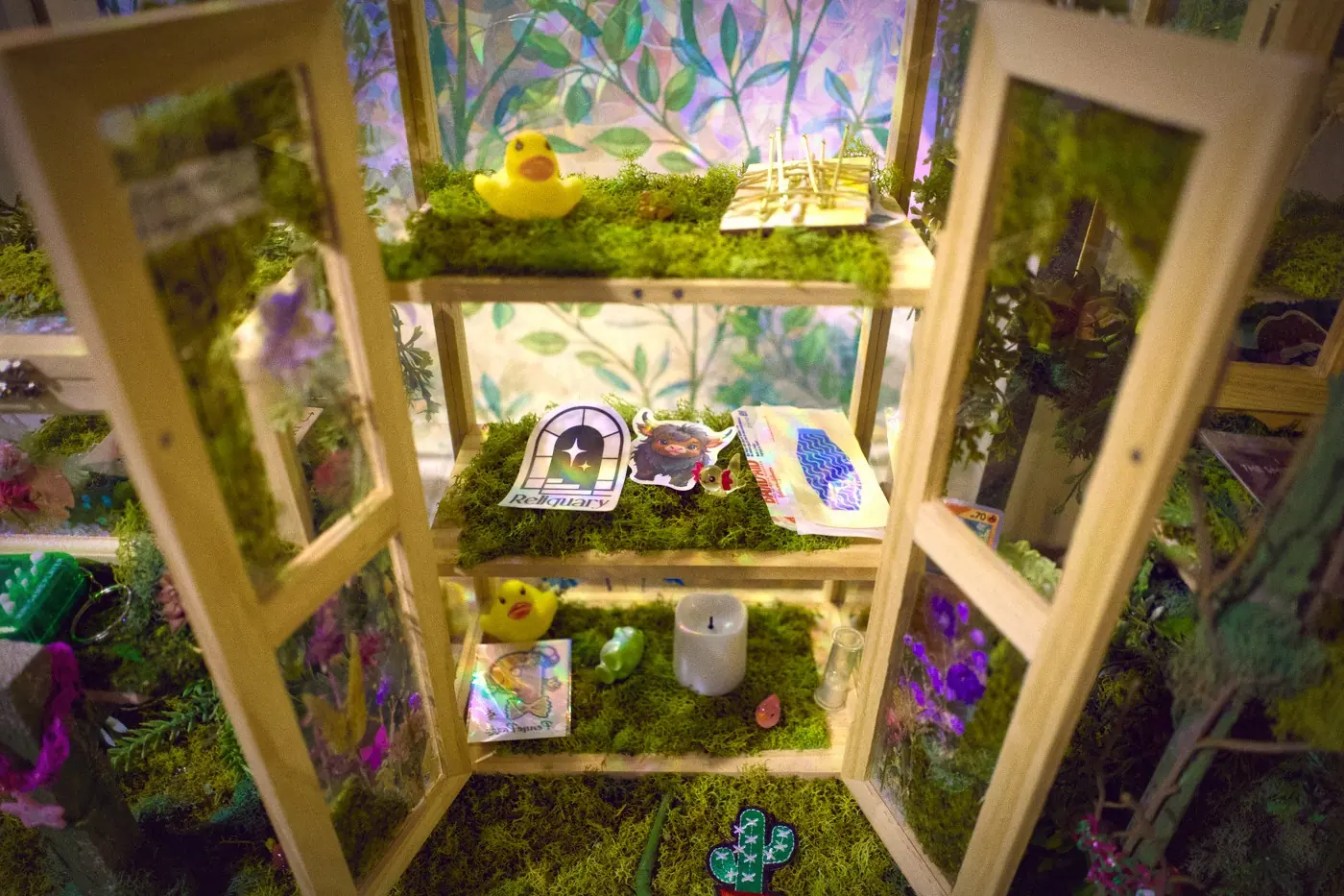 A small glass cabinet opened wide, its shelves lined with moss and holding trinkets left by visitors, a yellow rubber duck on the top shelf.